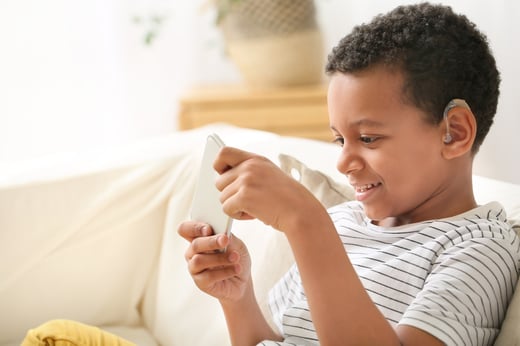 Little African-American boy with hearing aid using mobile phone at home